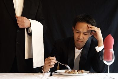 A stressed man eating food while another man stands besides him.